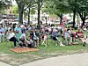 Crowd seated on chairs and blankets in a park lawn with trees and a walkway in the foreground.
