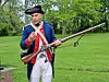 Man in an 18th-century-style uniform holding a musket on a grassy lawn.