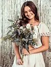Smiling woman in a white dress holding a bouquet of flowers against a weathered wooden wall.