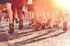 Low-angle view of several runners' legs and shoes on a sunlit road.