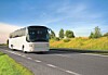 White coach bus driving on a highway beside grassy roadside and trees.