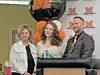 Three people stand beside a podium in front of an M-logo backdrop and balloons indoors.