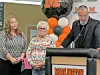 A man speaks at a podium decorated with balloons while two women stand beside him indoors.