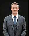 A man wearing a gray suit and blue tie stands against a black background.