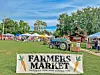 Farmers market booths and a tractor in a park with a Farmers Market sign.