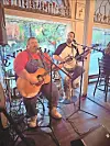 Two men play acoustic guitar and banjo while seated at microphones inside a restaurant.