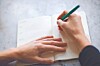 Hands writing in an open notebook with a green pen.