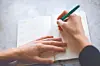 Hands writing in an open notebook with a green pen.