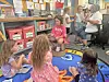 Children sit and stand in a classroom reading corner with colorful rugs, books, and storage bins.