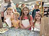 A group of children stands around a table holding notebooks and papers in a library.