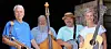 Four people posing outdoors with guitars and a bass in front of a wooden structure.