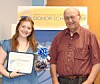 Two people stand indoors holding a scholarship plaque and a gift basket beside an award banner.