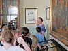 A guide points to framed artwork as visitors gather in an indoor gallery.