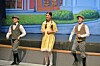 Three performers in period costumes stand on a stage before a painted backdrop.