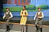 Three performers in period costumes stand on a stage before a painted backdrop.