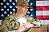U.S. soldier in camouflage uniform seated at a table with papers, in front of American flags.