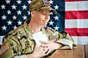 U.S. soldier in camouflage uniform seated at a table with papers, in front of American flags.