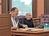 Two men seated at a wooden table in a wood-paneled room with papers, folders, and a water container.