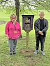 Two people standing beside a small wooden book exchange box in a grassy park-like setting.