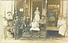 A group poses on the steps of a wooden storefront with porches and chairs.