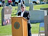 A man in a cap speaks at a podium outdoors in a cemetery with gravestones and flags behind him.