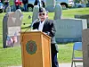 A man in a cap speaks at a podium outdoors in a cemetery with gravestones and flags behind him.