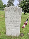 Weathered gravestone in grass with an American flag and a small marker nearby.