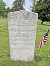 Weathered gravestone in grass with an American flag and a small marker nearby.