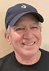 Smiling man wearing a dark baseball cap and black shirt in front of a beige wall.