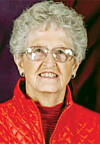 Portrait of an elderly woman wearing glasses and a red jacket against a dark background.