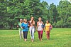 Five women walking on grass in a park, each carrying a tennis racket.