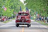 A vintage red fire truck with flags drives down a street as people watch from both sides.