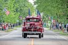 A vintage red fire truck with flags drives down a street as people watch from both sides.