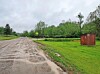 A muddy dirt road beside a grassy field with a small wooden shed and trees under cloudy skies.