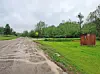 A muddy dirt road beside a grassy field with a small wooden shed and trees under cloudy skies.