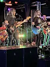 Live band performing on stage with guitars, drums, microphones and colorful lights.