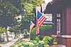 American flags displayed outside brick houses along a leafy residential street.