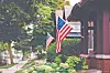 American flags displayed outside brick houses along a leafy residential street.