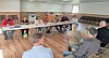 People seated around tables in a meeting room with chairs lined against the wall.