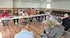 People seated around tables in a meeting room with chairs lined against the wall.