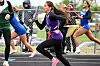 Track runners sprint in a relay race on an outdoor track beside a chain-link fence.