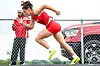 Athlete in red hurdles over a barrier as a coach stands nearby outside.