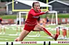 Athlete in red jumps over a hurdle on an outdoor track.
