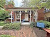 Red-brick historic house with a covered porch, arched supports, and a sign in the front yard.