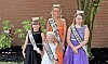 Four crowned pageant winners pose in formal dresses with sashes in front of a brick wall.