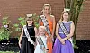Four crowned pageant winners pose in formal dresses with sashes in front of a brick wall.