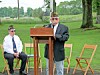 A man speaks at a podium outdoors as two uniformed veterans sit on folding chairs nearby.