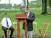 A man speaks at a podium outdoors as two uniformed veterans sit on folding chairs nearby.