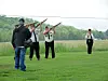 Several people stand in a field holding rifles, with one person in the foreground and others behind.