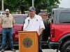 Man speaking at a wooden podium outdoors with two people and parked vehicles behind him.